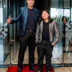 Two people in suits stand on a red carpet in front of glass doors, smiling at the camera.