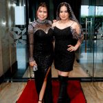 Two women in black evening dresses stand on a red carpet in a modern indoor setting.