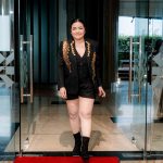 Person in a black outfit with gold details walks on a red carpet through glass doors.