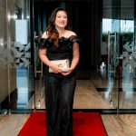 Woman in a black off-shoulder outfit holding a clutch stands on a red carpet indoors.