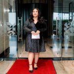 Woman in a black sequin dress stands on a red carpet in front of glass doors, holding a clutch.