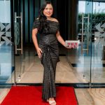 Woman in a sparkly black gown stands on a red carpet in front of glass doors.