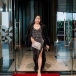 Woman in a black evening gown walking on a red carpet through glass doors.