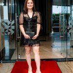 Woman in a black dress stands on a red carpet in front of glass doors, holding a clutch.