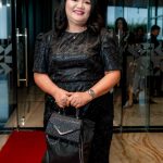 Woman in a black dress stands on a red carpet indoors, holding a black purse and smiling.