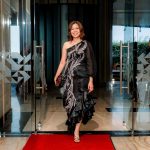 Woman in a black one-shoulder dress walks on a red carpet through glass doors.