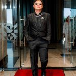 A man in a black suit and sunglasses walks on a red carpet through glass doors indoors.