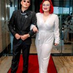 Two people in formal attire walk arm-in-arm on a red carpet in a modern indoor setting.