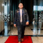 Man in a patterned suit walks on a red carpet through glass doors in a modern building.