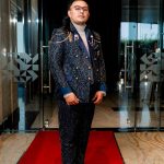 Man in a patterned suit and brown shoes standing on a red carpet in a modern hallway.