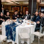 Seven people sit around a decorated table at a formal event in a banquet hall.