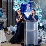 Two women stand at a podium speaking or singing at a decorated indoor event.