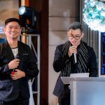 Two men in black suits speak at a podium during an indoor event with floral decor.