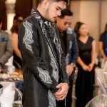 A man stands with his head bowed and hands clasped at a formal indoor event.