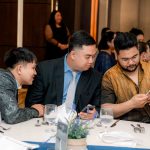 Three men sitting at a table, looking at a phone during a formal event.