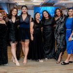Six women in black dresses and one woman in blue pose together at a formal indoor event.