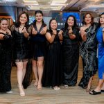 Seven women in formal dresses pose together at an indoor event, making hand heart gestures.