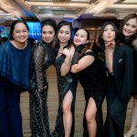 Six women in formal evening wear pose and smile together in a ballroom setting.