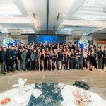 Large group of people pose together in formal attire in a bright, elegant banquet hall.