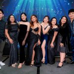 Eight people in formal attire pose together at an event with a "10th Anniversary" backdrop.