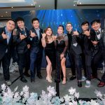 Eight people dressed formally pose and smile on a decorated stage at an event.