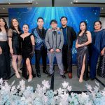 Nine people in formal attire pose together on a decorated stage with blue and white floral accents.