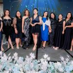 Ten women in formal black dresses pose together on a decorated stage with floral arrangements.