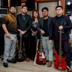 Five people pose indoors with electric guitars and a bass guitar, standing in front of music equipment.