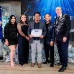 Five people pose indoors; the center person holds a certificate. Festive blue decor behind them.