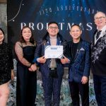 Five people pose at an event; one holds a certificate in front of an anniversary backdrop.