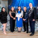 Five people pose indoors; one woman holds a certificate at an anniversary event.