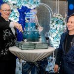 Two men in suits prepare to cut a blue anniversary cake at a decorated event.