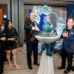 Four people in formal attire stand around a table with a decorative cake at an indoor event.
