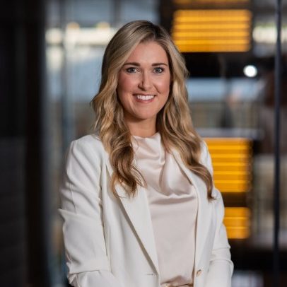Woman with long blonde hair in a white blazer at Leaders Conference 2025, smiling indoors.