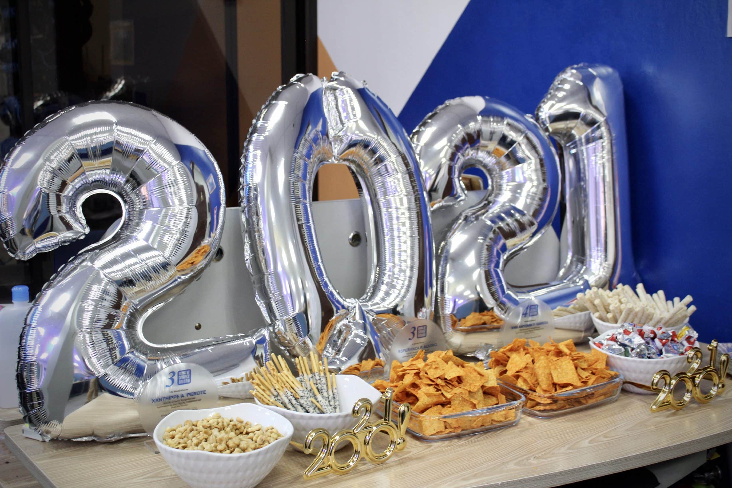 A table with silver "2021" balloons, several bowls of snacks including chips and nuts, and decorative "2021" items set up for a year end party celebration.