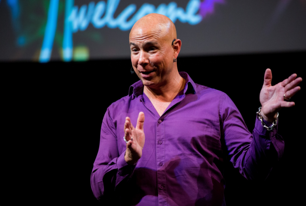 A bald man in a purple shirt gestures on stage at Leaders Conference 2025, "welcome" behind him.