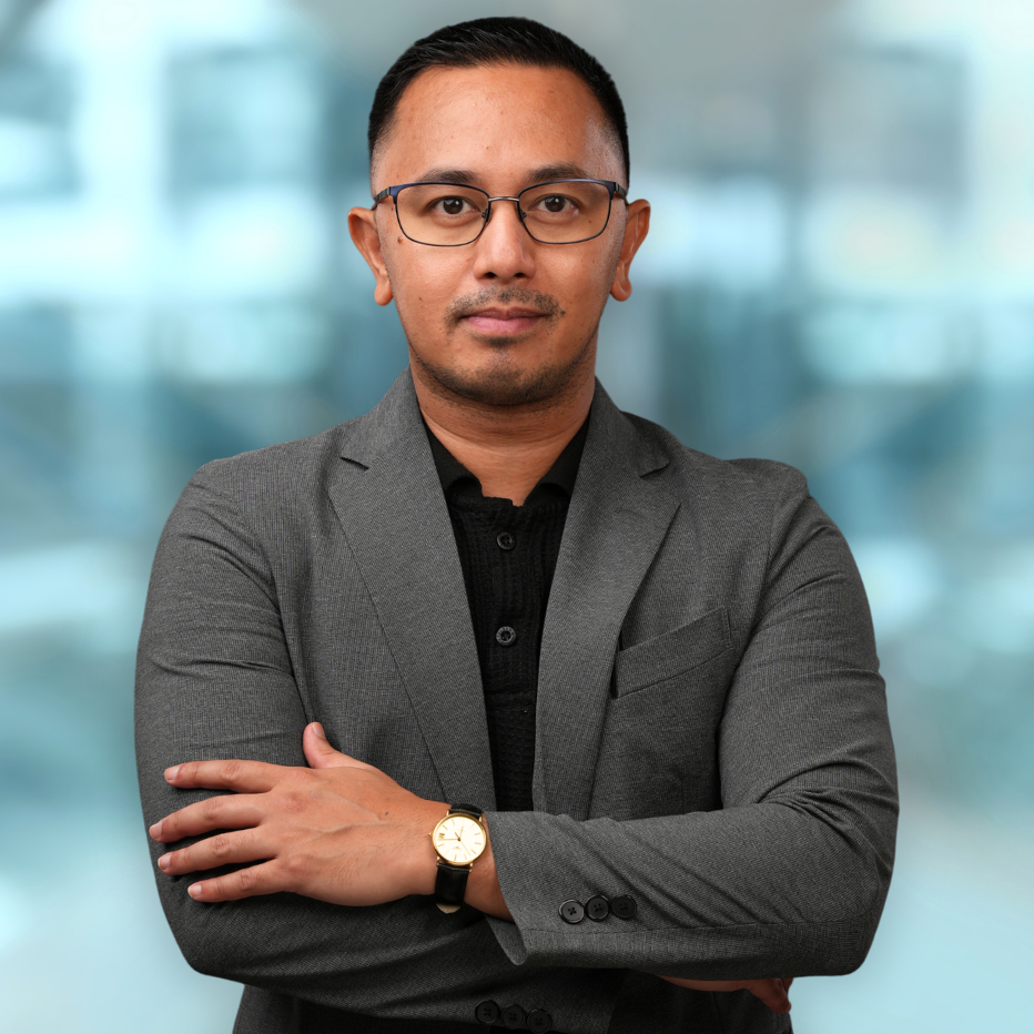 A man in a gray suit and glasses stands with arms crossed against a blurred background.