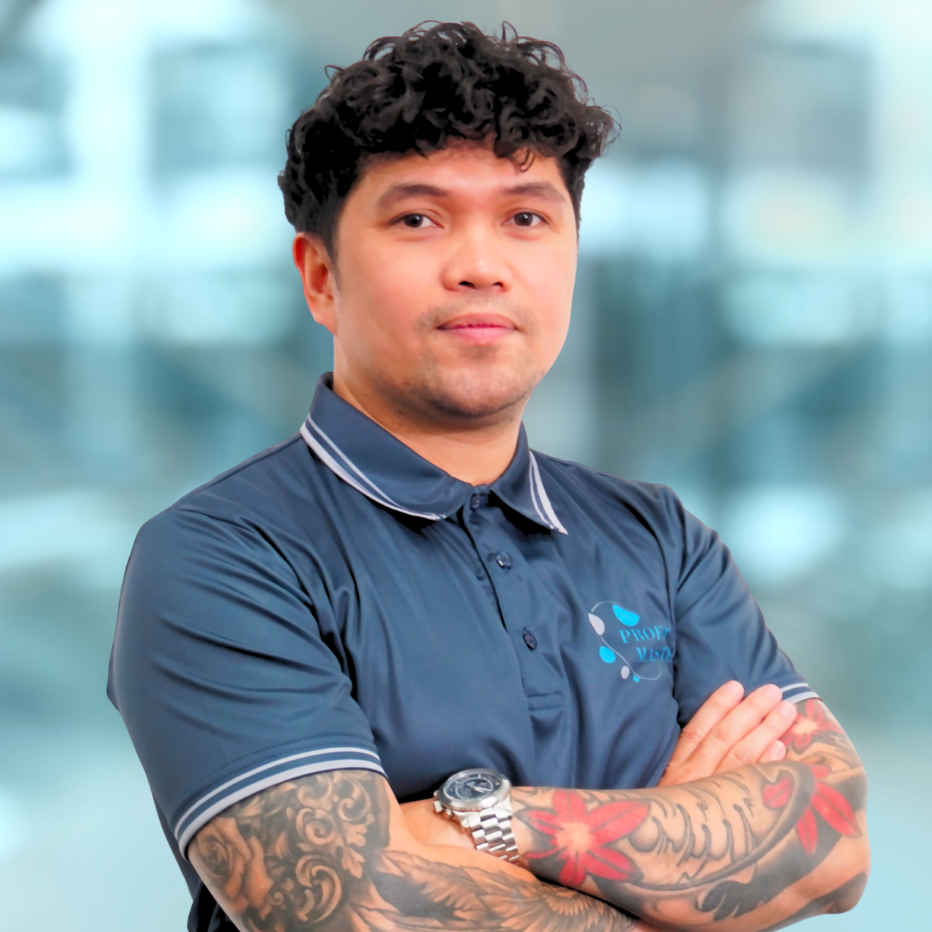Man with tattooed arms in a blue collared shirt stands with arms crossed, smiling slightly.