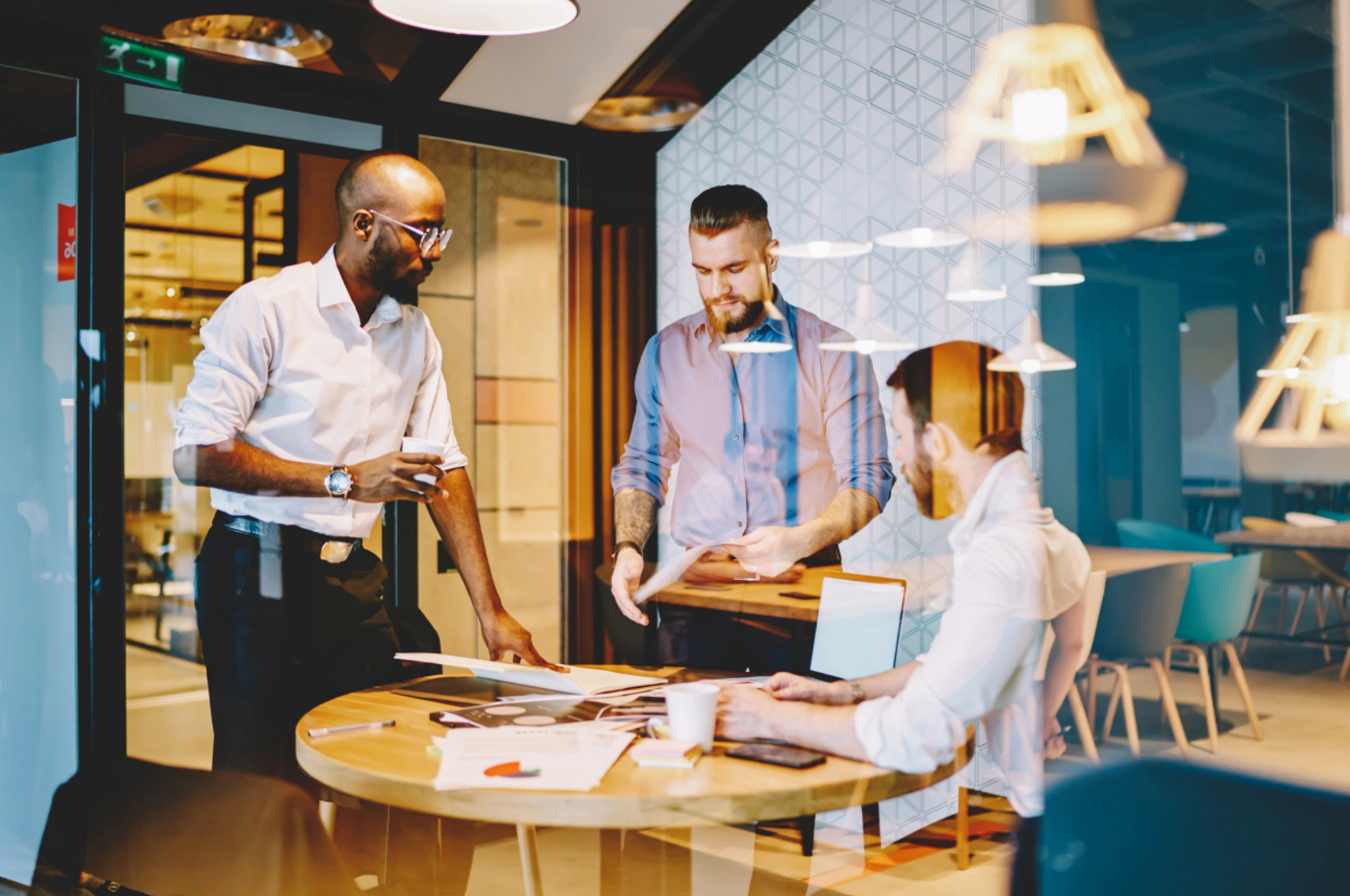 Three men discuss documents around a round table during a Lunch & Learn in a modern office.