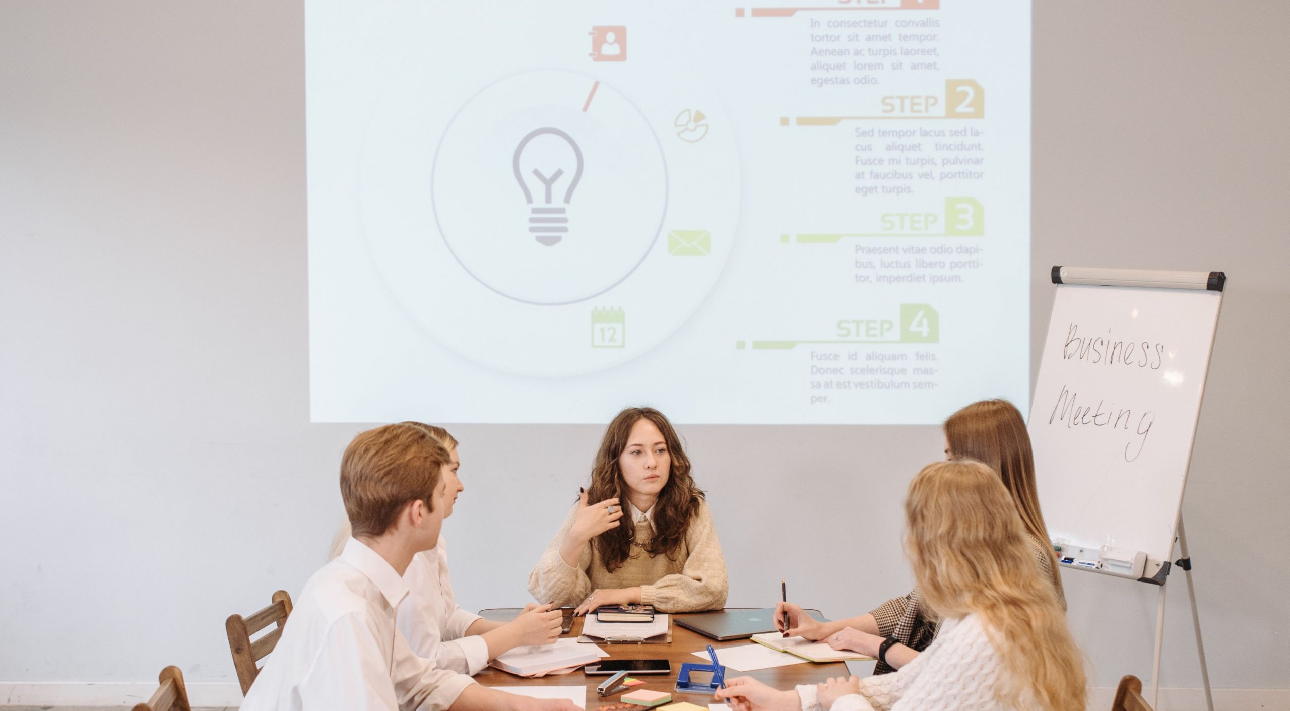 A group of people sits around a table having a discussion, with a presentation projected on the wall and a flip chart with "How to Create Great Content" written on it.